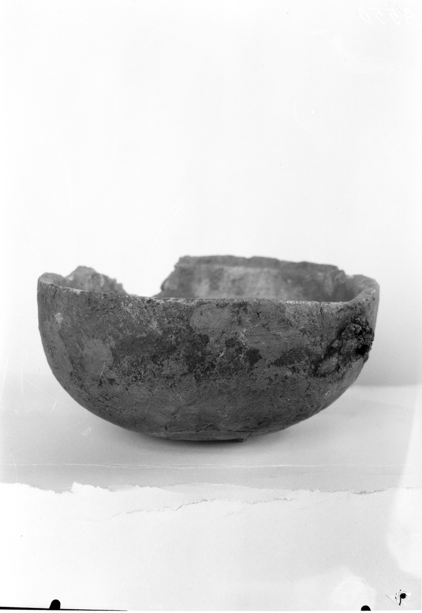 "4946 a. Gryte av klebersten av den vanlige type R. 729, tverrmålet i munningen 20,3 cm., høiden ca. 9 1/2 cm. Omtrent fullstendig bevart, bare en del av overkanten mangler på den ene side. Rester av jernhanken foreligger også."
