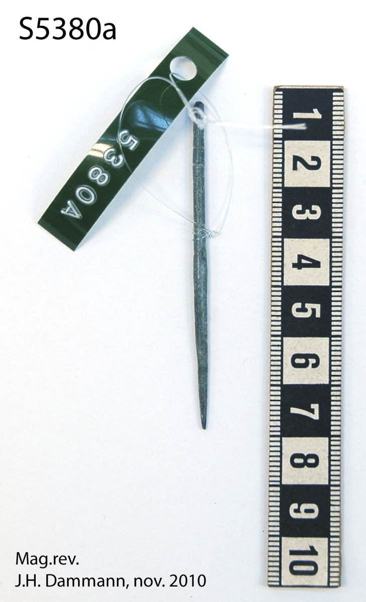 "Tynn nål av bronse, 6,6 cm.l., 5 omløpende furer innrisset om nålens øvre del." Ny beskrivelse etter revisjon: Nål av bronse i samsvar med beskrivelsen. Odden er intakt. De innrissede linjene er meget enkelt utført. Nålen er tykkest på midten, diam.: 2,4 mm.