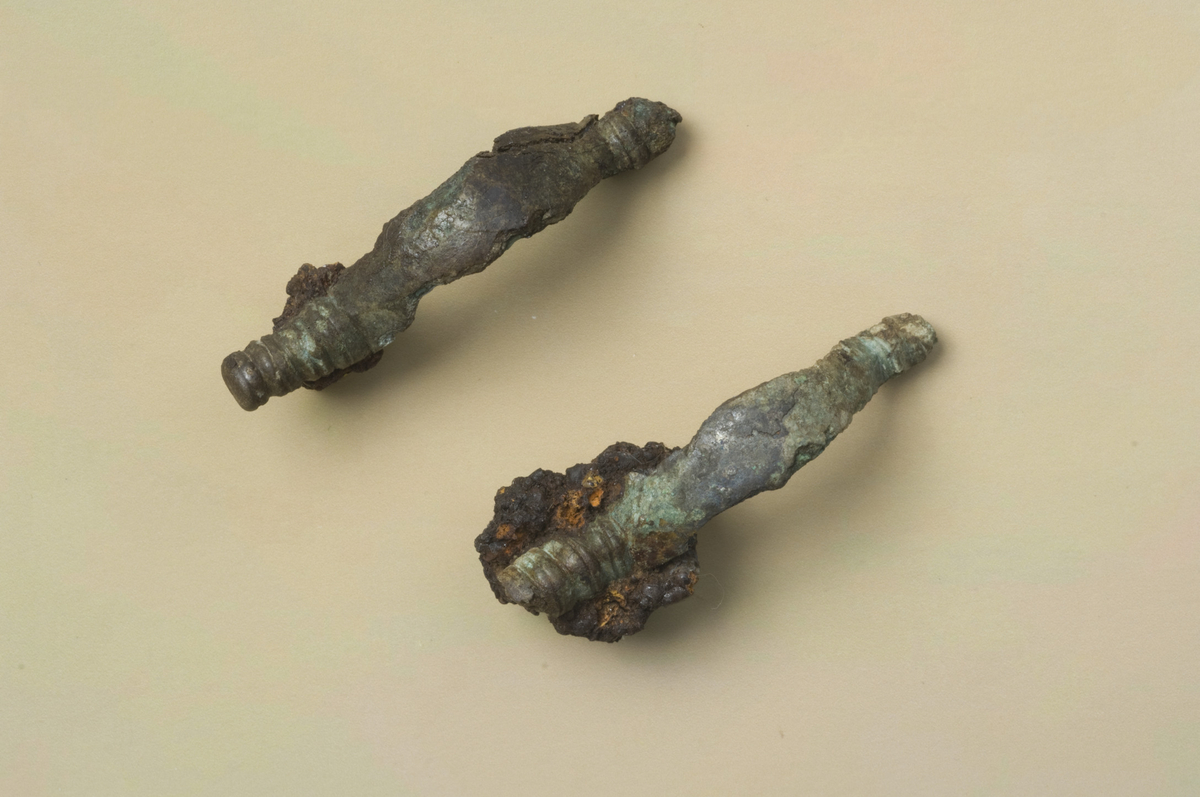 " To identiske likearmete bronsespenner av typen Shetelig: Smaa spænder fra f.v.t fig. 69, s. 85. Begge er delvis ødelagt av stein i gravkammeret, men de har sannsynligvis bare hatt fire tverrgående vulsster som avslutning på endene og ikke som fig. 69, fem tverrgående vulster og en endeknopp. De har hatt jernnål. L: ca 50 mm, stbr: 10 mm." Ny beskrivelse etter revisjon: To likearmede spenner, i samsvar med beskrivelsen. Spenne fnr. 10 har alle fire vulster bevart i ene enden, l.: 46 mm, stbr.: 9 mm. Nålspiralen bevart som et 11 x 11 mm stor korrosjonsklump. Spenne fnr. 5 er skadet i begge ender, l.: 43 mm, stbr.: 9 mm. Nålspiralen er bevart som en 20 x 17 mm stor korrosjonsklump.Tre små frag. mineralisert tekstil ligger løst.
