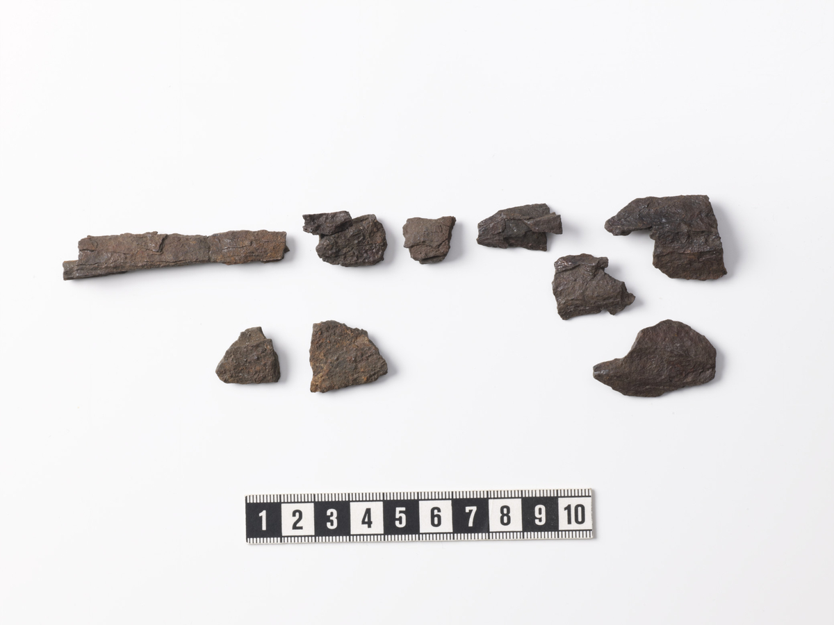 8092 (e) Ni fragmenter av lansespiss. (1) Fragment av hals, store deler avsprengt, med utside krum i bredderetningen. Stl: 65 mm, stbr: 13 mm.  (2) Fire fragmenter av fal med mineralisert tekstil på falens innside, stl: 23-32 mm. Ett fragment er limt av to biter. Det lengste fragmentet har fasettert omkrets, stdiam: ca 32 mm. (3) Fragment av fal uten tekstil på innsiden. Fragmentet har fasettert omkrets, stl: 35 mm. (4) Fragment av hals (?), stl: 15 mm. (5) To tresidige fragment av blad med egg, stl: hhv. 18 og 23 mm, stbr: hhv. 16 og 20 mm.
/Skilt ut fra underbokstav c./