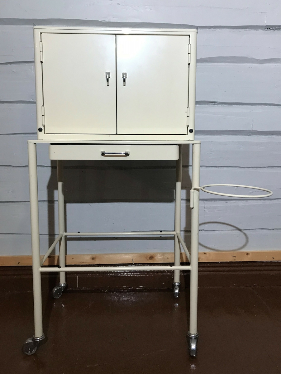 Bord på hjul, med skuff og plass til glasshylle under. Skap som er fastmontert oppå bordet. Skapet har plass for hyller inni og det er hyller i dørene. Det er montert en metallring på ene benet til bordet. 
