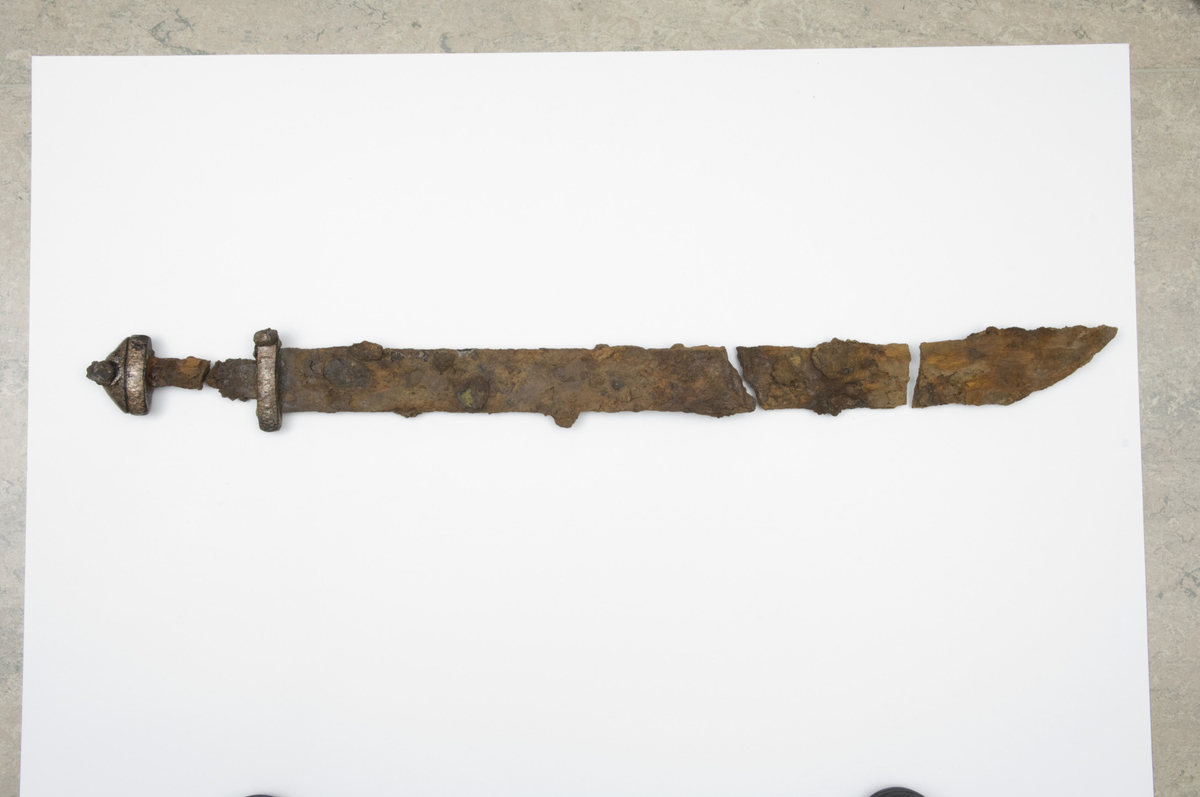 b) Sverd av jern. Type H, enegget, i fire deler. Knapp, overhjalt og øvre del av grep l. 96 mm. Klingen i tre deler som passer sammen. Det lengste fragmentet med nedre del av grep og underhjalt l. 498 mm. Klingelengde 751 m, br. 59 mm like under underhjaltet. Grepet delt i to, bevarte l. 55 og 52 mm, br. 26 mm. Underhjalt l. 94 mm, h. 19 mm, t. 31 mm. Overhjalt l. 75 mm, h. 16 mm. Knapp h. 29 mm, t. 32 mm. Samlet høyde knapp, overhjalt og korrosjonsblære på knapp 56 mm. Hjalter med elliptisk tverrsnitt. Ei nagle synlig på røntgen mellom overhjalt og knapp. Hjalter og knapp dekorert i trappemønster av tynne plater av sølv, kobber/bronse. Messingplate påfestet under overhjalt. (Fnr. 2410.) 