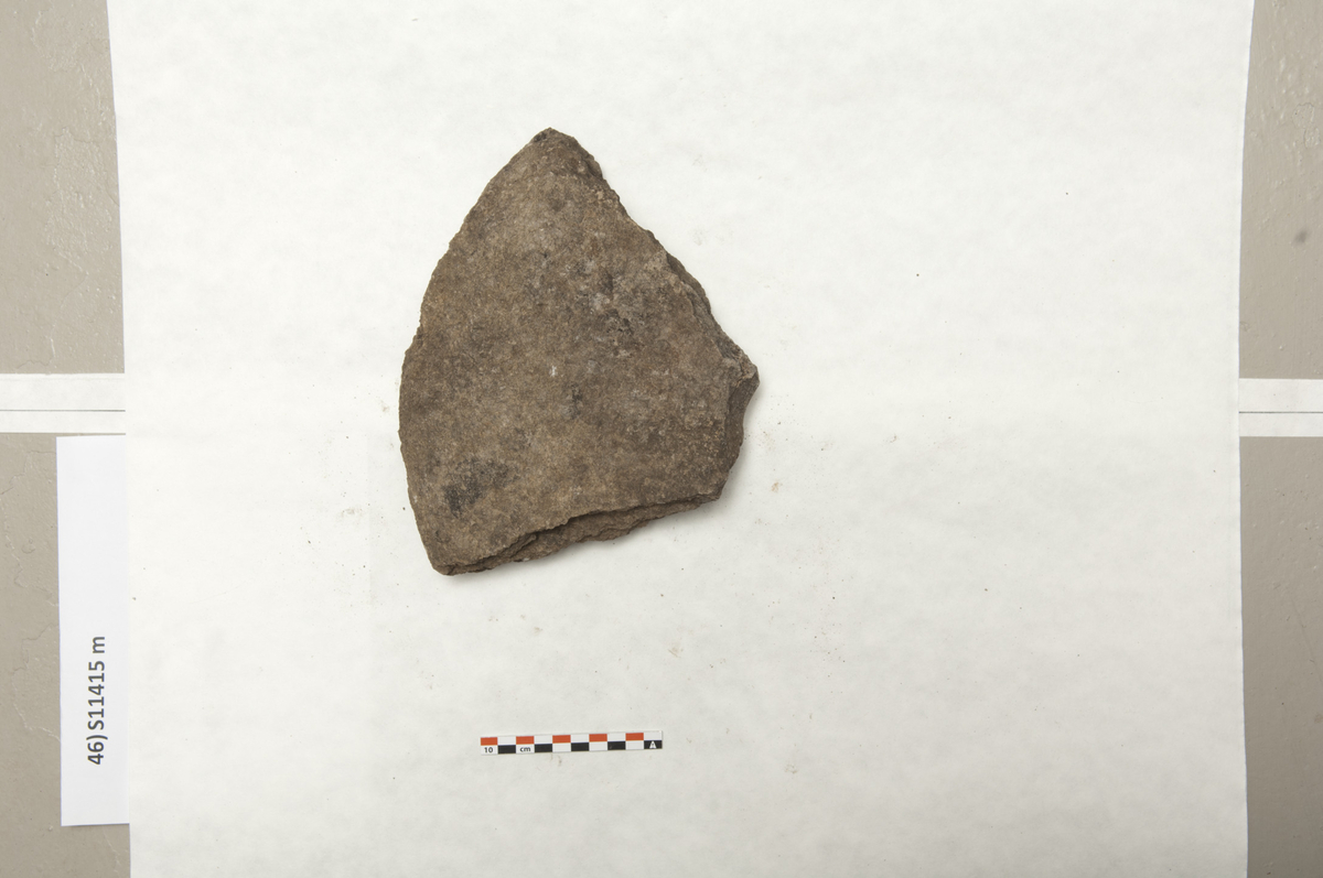 Et fragment av drejekvern med trapezoid form, med flat underside og svakt buet overside. På den korteste side ses profil av midthullet. St.m: 260 mm, vekt: 2712,2 g. (Fnr. 384)