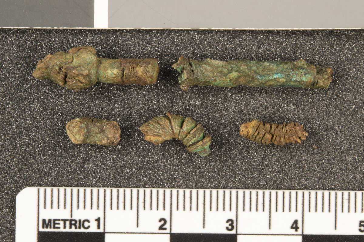 2 fragmenter i bronse av noe som sannsynligvis er en pinsett. Fragmentene er svært dårlig bevart og var til konservering da katalogiseringen ble gjort.
a) Ett 3,6 cm lang fragment som er svakt buet i enden. Bruddflater i begge endene. Kan muligens være den ene armen på en pinsett. b) Ett 2,7 cm langt fragment med en klump i enden som kan være grepet/hodet på pinsetten. Begge fragmentene er 0,4 cm brede med rundt tverrsnitt.