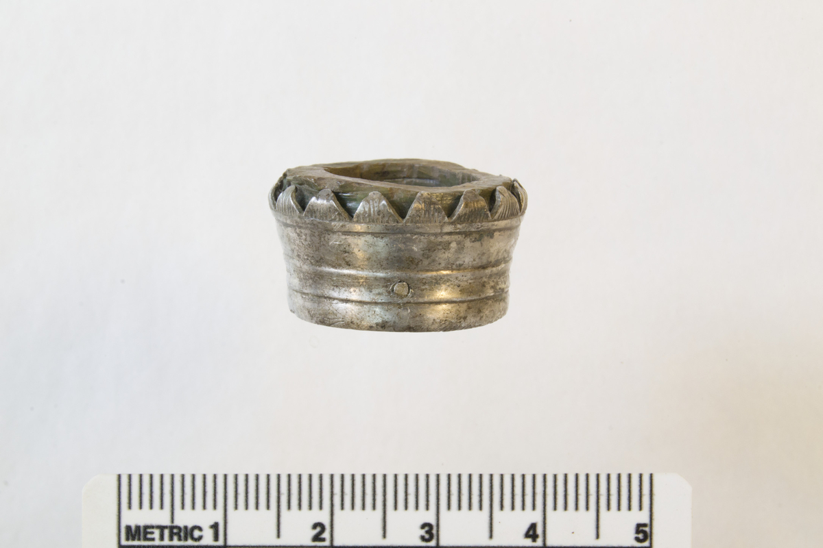 Profilert beslag/holk til kniv av sølvlegering med endeskive og to små spiker til festing. St. diam.: 24 mm, minste diam.: 21 mm, l.: 18 mm. Beslaget har seksten små riflede trekanter med spissene delvis krummet over endeskiven. Endeskiven er av gevir (?), t.: 4 mm, br.: 4 mm. Inni beslaget sitter et tynt, sirkulært trespon. 