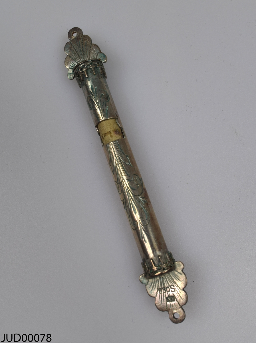 Mezuza tillverkad av silver, i en rund design och trekantigt, platt avslut. Graverad snirklig dekor. Textrulle finns inuti.
