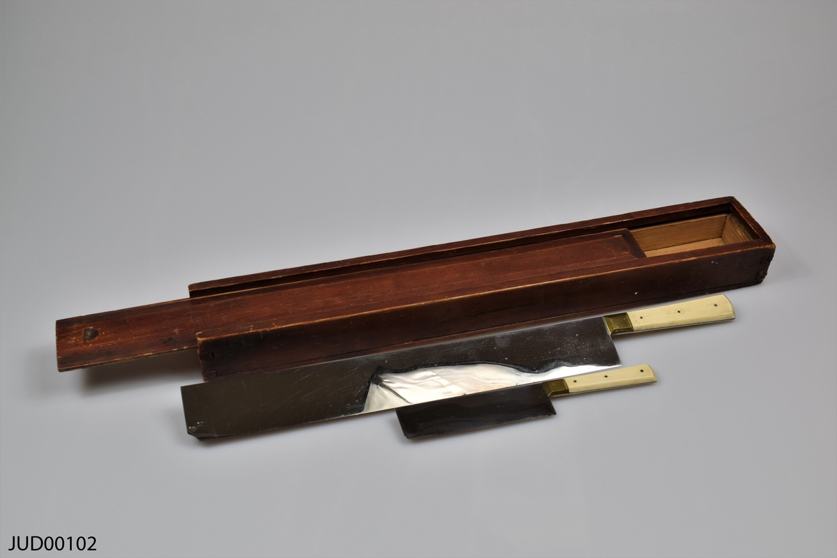 Rektangulär smal trälåda innehållandes två knivar. Knivarna har vita handtag och rektangulära blad.
