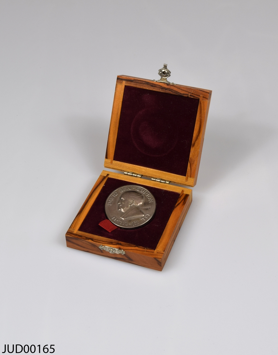 Bronsmedalj med tillhörande träetui, klätt invändigt med röd sammet. Lås på etuiets framsida, tillverkad av metall. Medaljen är dekorerad med text på fram- och baksidan, samt en läsande arbetare på ena sidan och en man i profil (David Den Gurion) på andra sidan.
