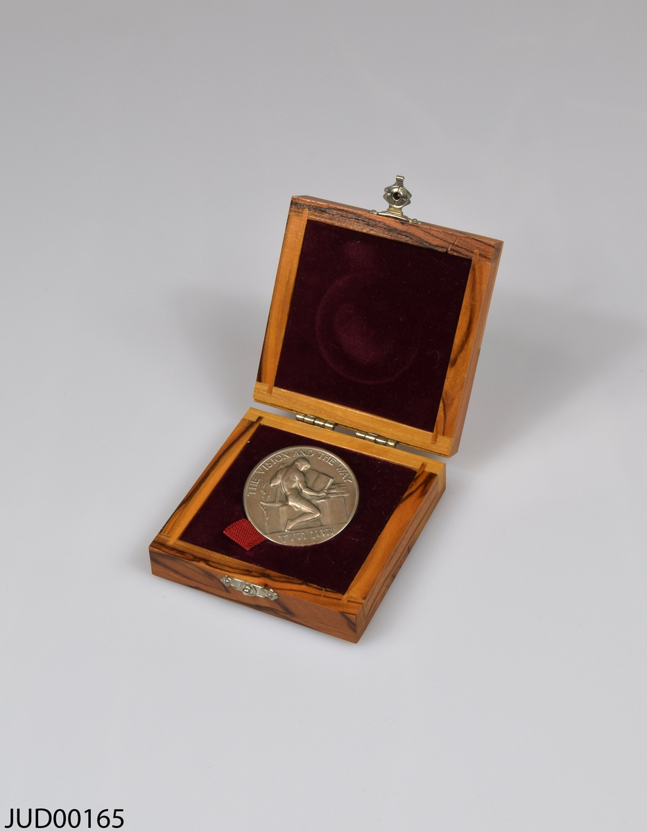 Bronsmedalj med tillhörande träetui, klätt invändigt med röd sammet. Lås på etuiets framsida, tillverkad av metall. Medaljen är dekorerad med text på fram- och baksidan, samt en läsande arbetare på ena sidan och en man i profil (David Den Gurion) på andra sidan.