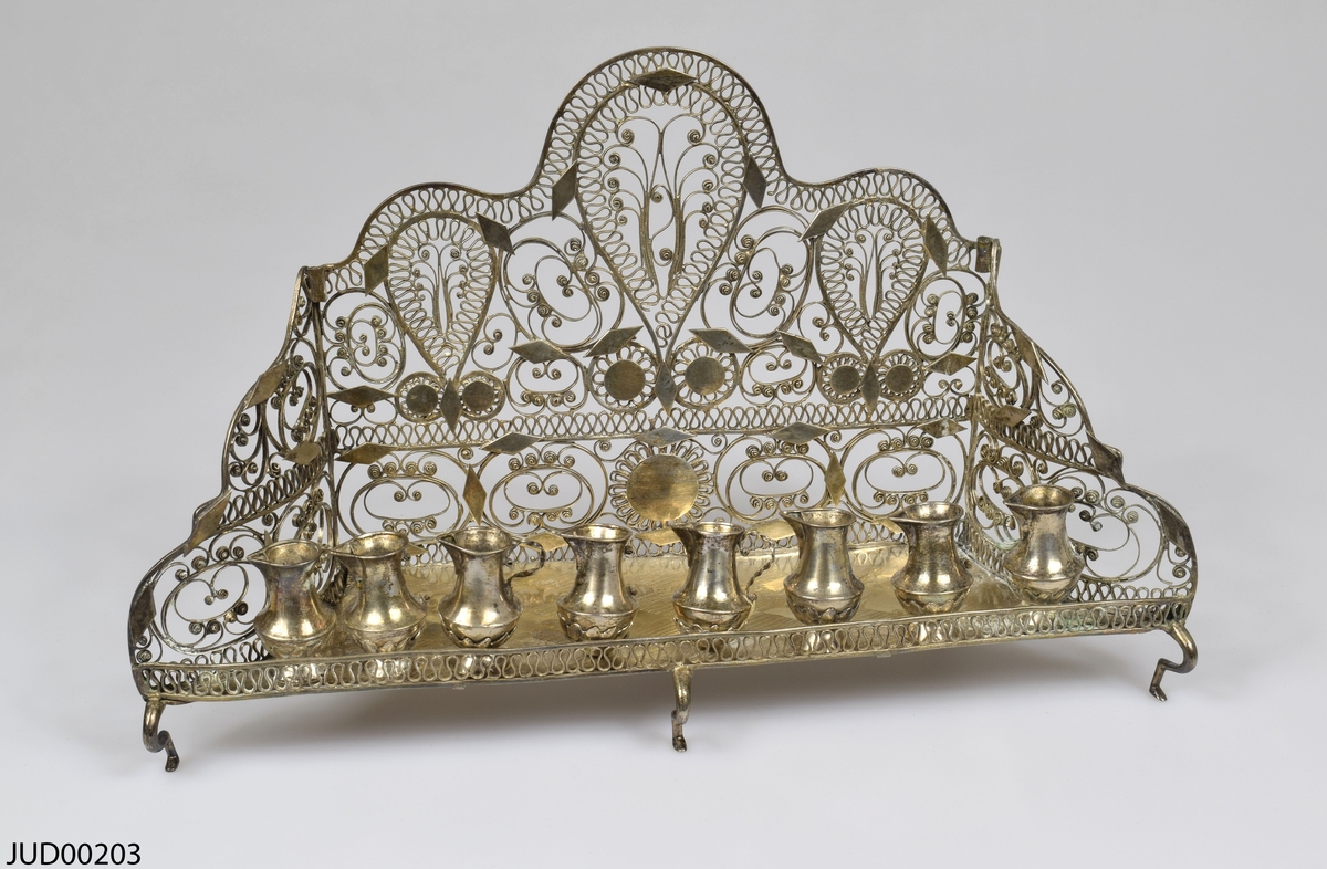 Chanukia tillverkad av silver. Filigranarbete med blomstermönster. Även dekorerad med utskurna romber och cirklar fastlödade ovanpå filigranen. Åtta ljushållare i form av kannor, som är fastskruvade i bottenplattan. Ristad rombdekor på bottenstycket bakom kannorna. Två otydliga stämplar på bottenplattan, bakom kannorna.