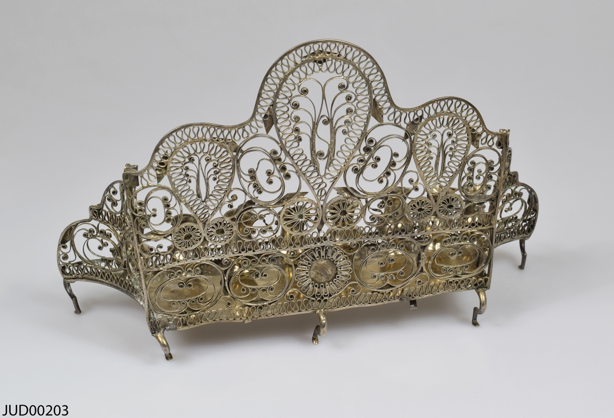 Chanukia tillverkad av silver. Filigranarbete med blomstermönster. Även dekorerad med utskurna romber och cirklar fastlödade ovanpå filigranen. Åtta ljushållare i form av kannor, som är fastskruvade i bottenplattan. Ristad rombdekor på bottenstycket bakom kannorna. Två otydliga stämplar på bottenplattan, bakom kannorna.