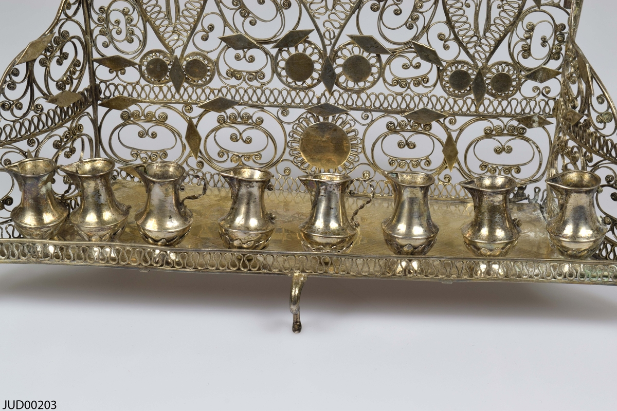 Chanukia tillverkad av silver. Filigranarbete med blomstermönster. Även dekorerad med utskurna romber och cirklar fastlödade ovanpå filigranen. Åtta ljushållare i form av kannor, som är fastskruvade i bottenplattan. Ristad rombdekor på bottenstycket bakom kannorna. Två otydliga stämplar på bottenplattan, bakom kannorna.