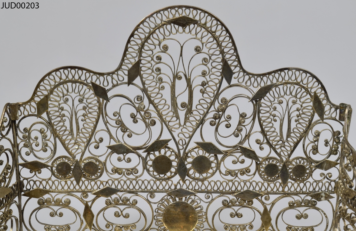 Chanukia tillverkad av silver. Filigranarbete med blomstermönster. Även dekorerad med utskurna romber och cirklar fastlödade ovanpå filigranen. Åtta ljushållare i form av kannor, som är fastskruvade i bottenplattan. Ristad rombdekor på bottenstycket bakom kannorna. Två otydliga stämplar på bottenplattan, bakom kannorna.