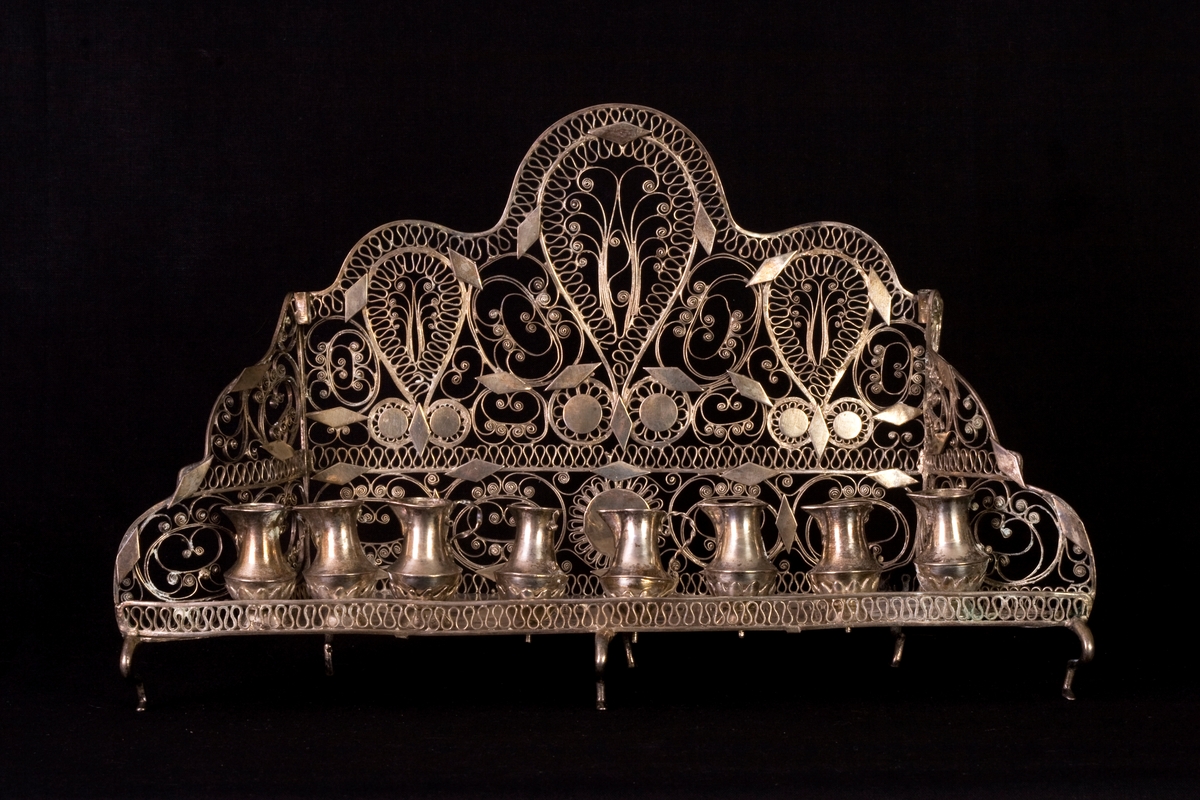 Chanukia tillverkad av silver. Filigranarbete med blomstermönster. Även dekorerad med utskurna romber och cirklar fastlödade ovanpå filigranen. Åtta ljushållare i form av kannor, som är fastskruvade i bottenplattan. Ristad rombdekor på bottenstycket bakom kannorna. Två otydliga stämplar på bottenplattan, bakom kannorna.