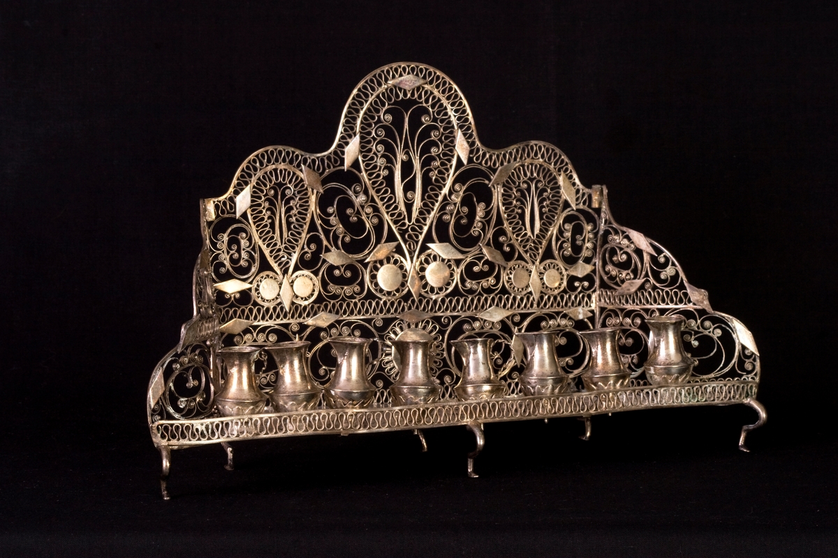 Chanukia tillverkad av silver. Filigranarbete med blomstermönster. Även dekorerad med utskurna romber och cirklar fastlödade ovanpå filigranen. Åtta ljushållare i form av kannor, som är fastskruvade i bottenplattan. Ristad rombdekor på bottenstycket bakom kannorna. Två otydliga stämplar på bottenplattan, bakom kannorna.