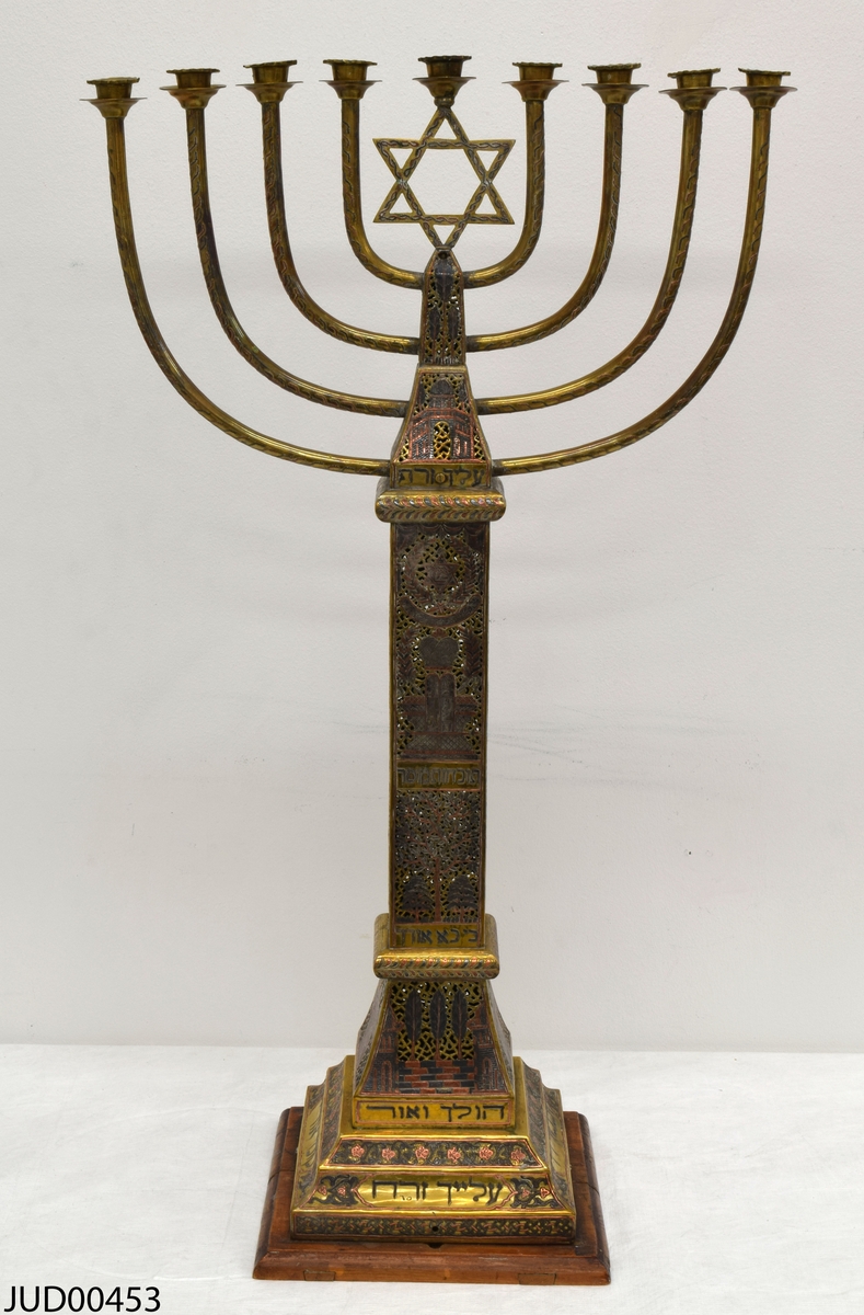 Stor chanukkaljusstake tillverkad av mässing, i form av en obelisk dekorerad genombruten dekor i rött och svart. Davidsstjärna på toppen mellan ljushållarna.