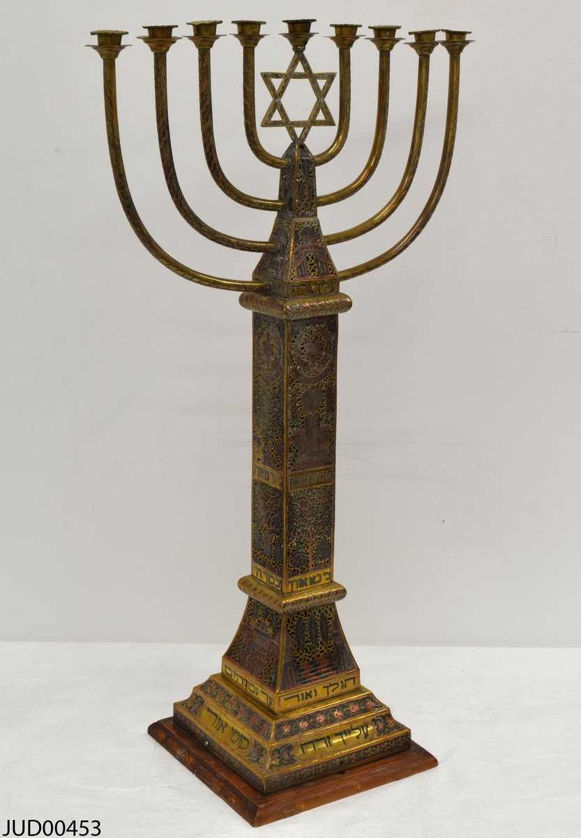 Stor chanukkaljusstake tillverkad av mässing, i form av en obelisk dekorerad genombruten dekor i rött och svart. Davidsstjärna på toppen mellan ljushållarna.