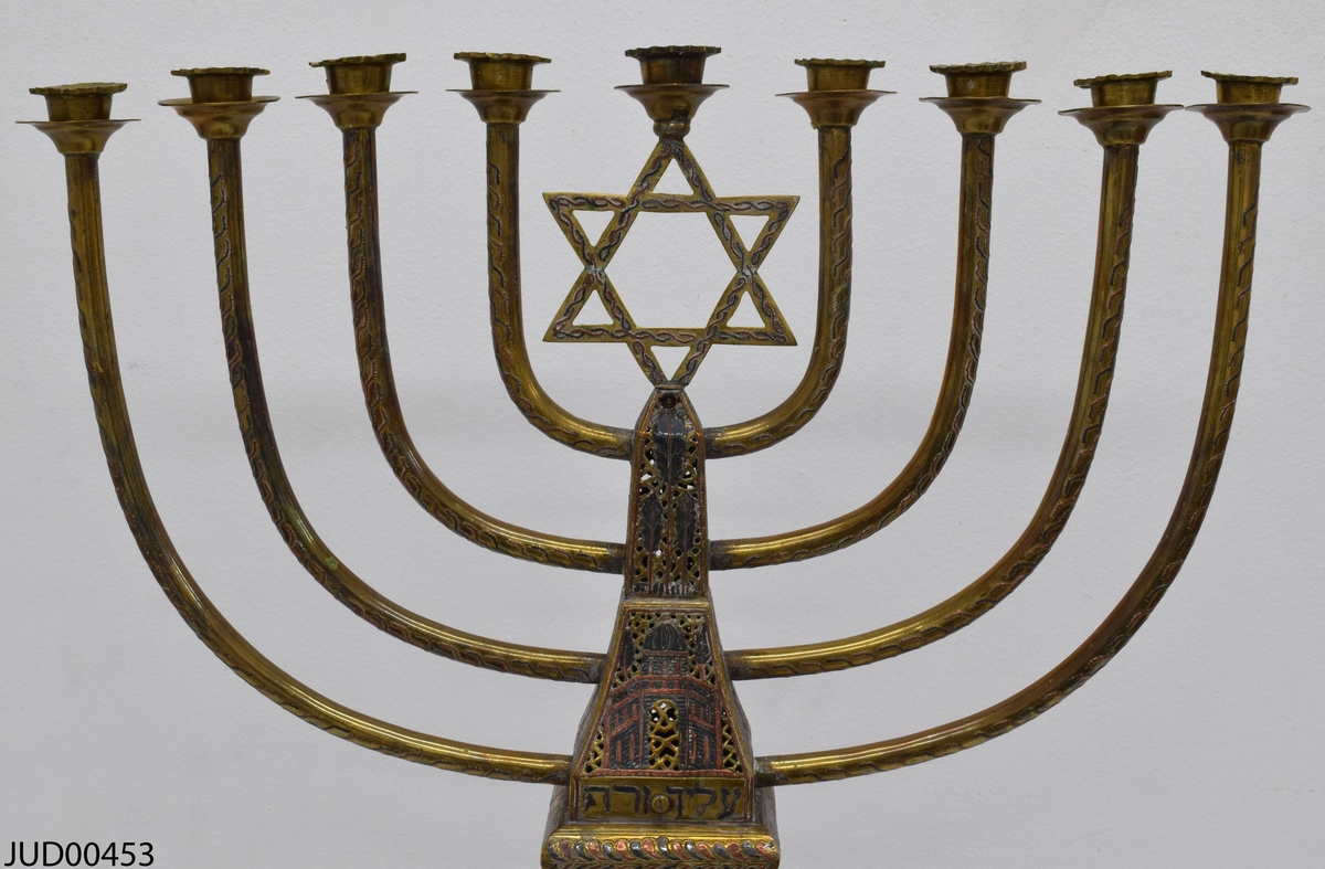 Stor chanukkaljusstake tillverkad av mässing, i form av en obelisk dekorerad genombruten dekor i rött och svart. Davidsstjärna på toppen mellan ljushållarna.