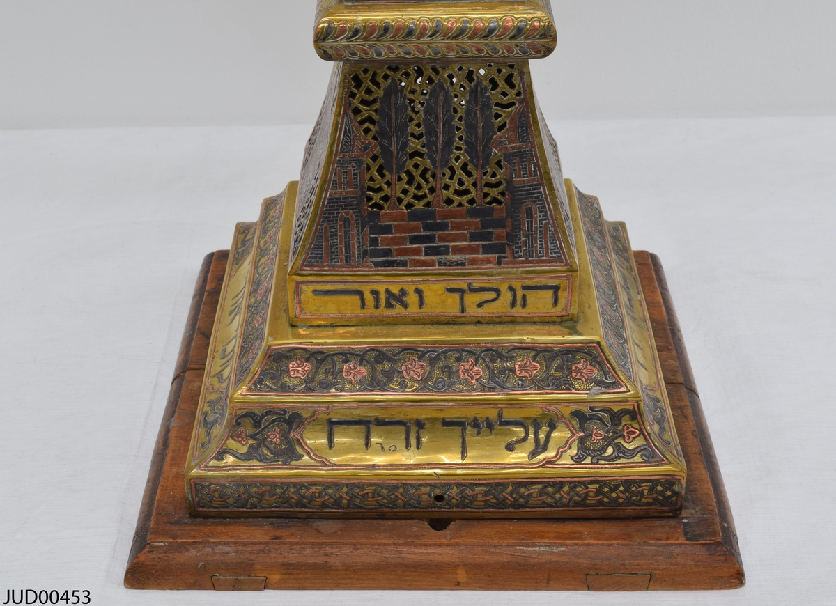 Stor chanukkaljusstake tillverkad av mässing, i form av en obelisk dekorerad genombruten dekor i rött och svart. Davidsstjärna på toppen mellan ljushållarna.