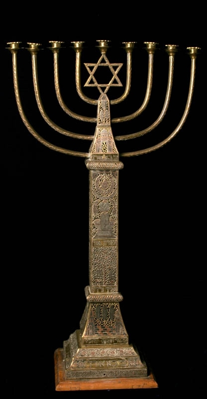 Stor chanukkaljusstake tillverkad av mässing, i form av en obelisk dekorerad genombruten dekor i rött och svart. Davidsstjärna på toppen mellan ljushållarna.