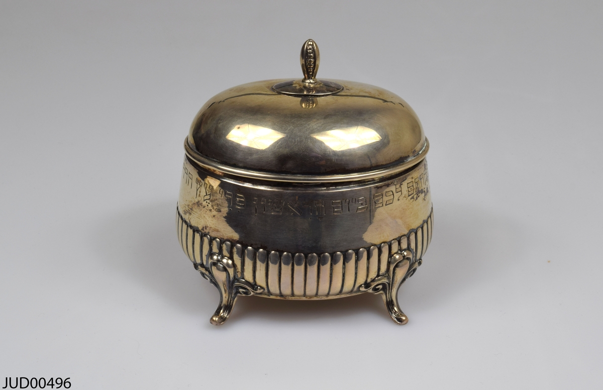 Silverask, med lock fastsatt på gångjärn. Står på fyra korta ben. Enkelt dekorerad med ristad hebreisk text.