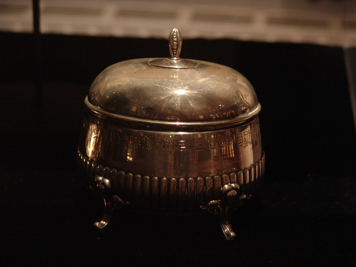Silverask, med lock fastsatt på gångjärn. Står på fyra korta ben. Enkelt dekorerad med ristad hebreisk text.
