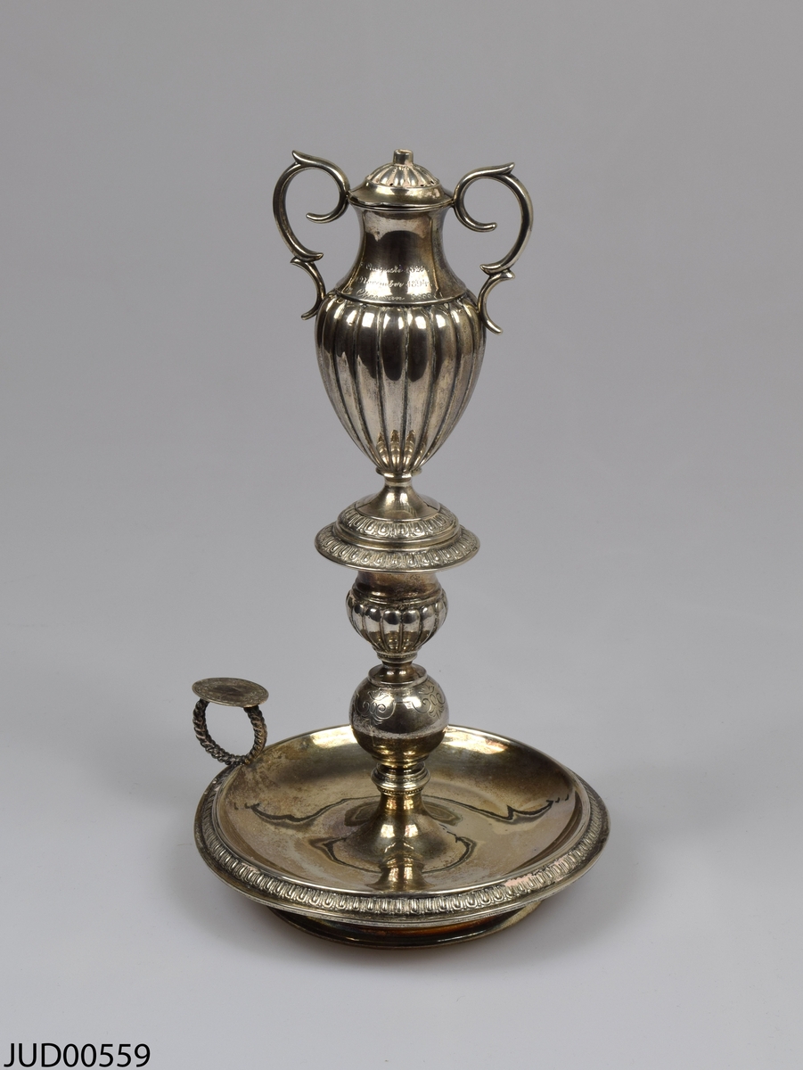 Årslampa. Svensk silverljusstake i tre delar. Toppen är i form av en amfora med handtag och lock. Ristade årtal på amforan - 1829, 1894 samt 5655.