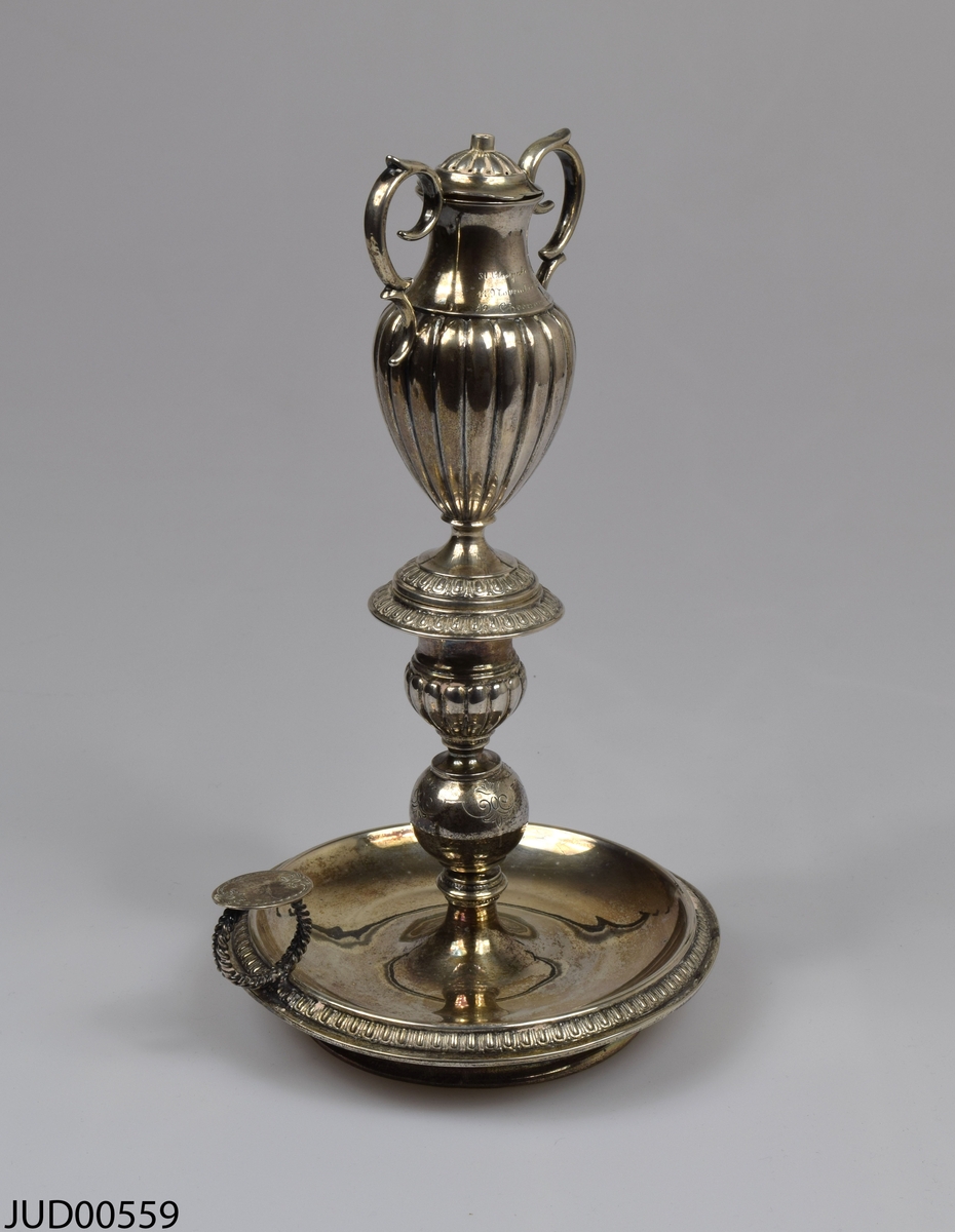 Årslampa. Svensk silverljusstake i tre delar. Toppen är i form av en amfora med handtag och lock. Ristade årtal på amforan - 1829, 1894 samt 5655.