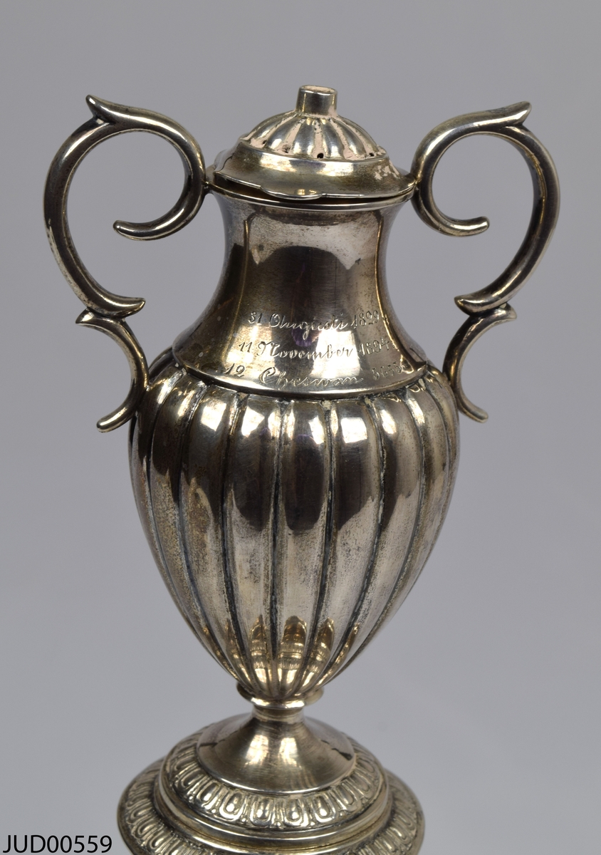 Årslampa. Svensk silverljusstake i tre delar. Toppen är i form av en amfora med handtag och lock. Ristade årtal på amforan - 1829, 1894 samt 5655.