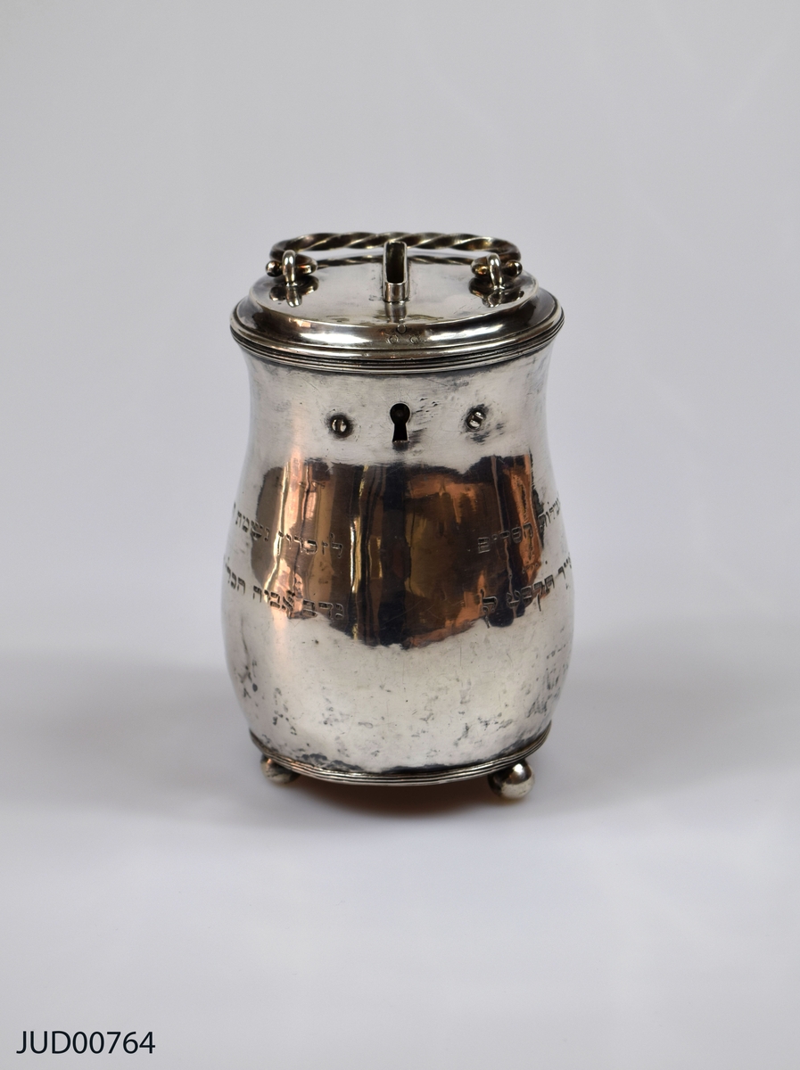 Insamlingsbössa från 1809, tillverkad av silver. Myntinkast ovanpå locket. Även vridet handtag fastsatt i locket. Nyckelhål precis nedanför lock. Inskription på hebreiska runt om. Står på tre kulfötter.