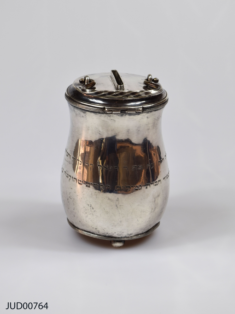 Insamlingsbössa från 1809, tillverkad av silver. Myntinkast ovanpå locket. Även vridet handtag fastsatt i locket. Nyckelhål precis nedanför lock. Inskription på hebreiska runt om. Står på tre kulfötter.