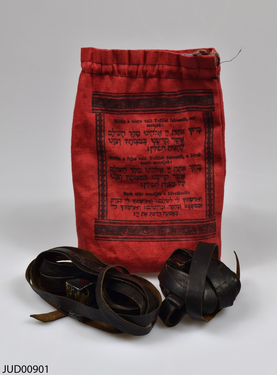 Ett par tefillin (böneremmar) med tillhörande påse. Kapslarna är tillverkade av trä, medan remmarna är tillverkade av svart läder. Påsen är tillverkad av rött tyg, och påsens insida är klädd med lilafärgat brokad. Påsen är dekorerad med svart hebreisk och ungersk text tryckt på ena sidan, medan påsens andra sida är dekorerad med stentavlorna tryckta i svart.