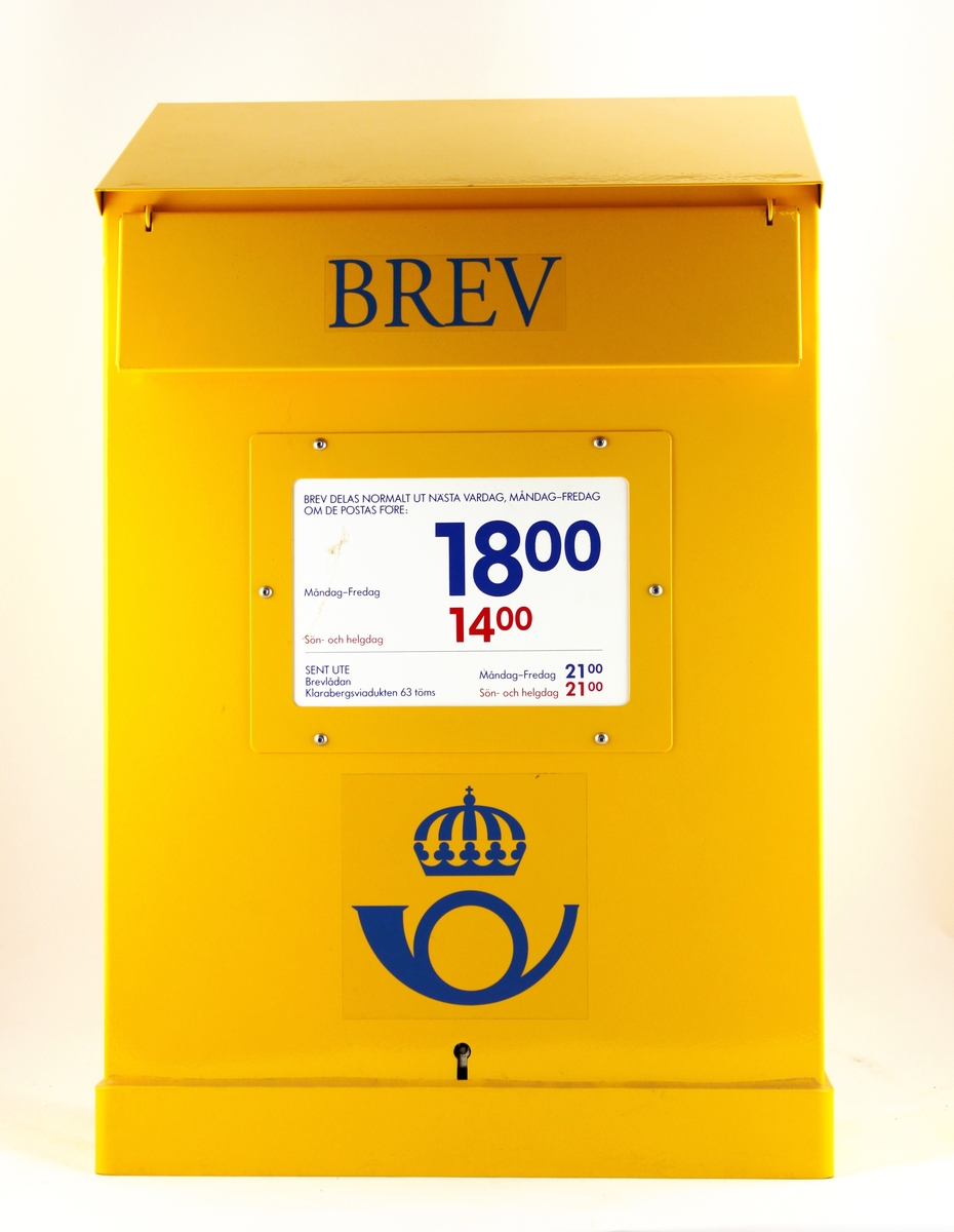 Gul brevlåda av vanlig modell med blå text på klaffen. En postsymbol i blått under anslagsfönstret. Anslag om tömningstider sitter i anslagsfönstret. Nyckel tillhör.
