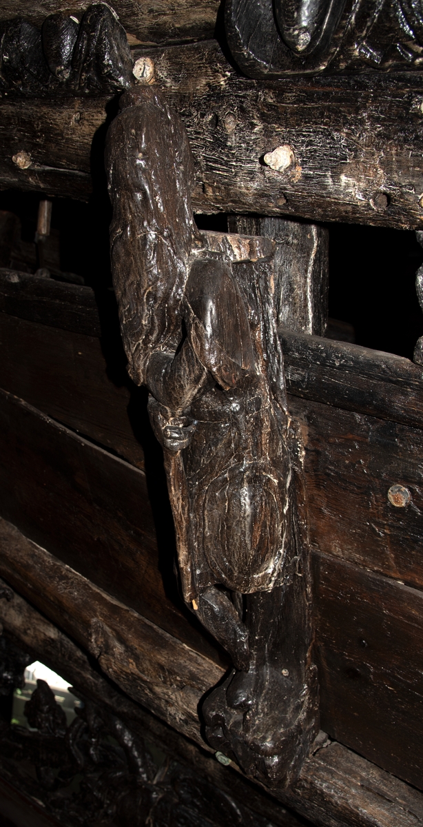 Skulptur föreställande en mansfigur med långt hår och ett stort böljande patriarkskägg. Pannan är rynkad. På huvudet sitter en stor, slät hjälm med långa, nedhängande plymer. Figuren är iförd en enkel dräkt, en rustning av thoraxmodell eller möjligen en civil dräkt. Vid vänster sida hänger från bältet en oval sköld med bred kant. Figuren stödjer sig mot en stav.



Figurens huvud är kraftigt vridet åt vänster medan bålen, främst dess övre del, är vriden något åt höger. Benen är frontalställda och korslagda. Kroppstyngden vilar helt på det vänstra benet. Det högra benet är placerat framför det vänstra. Figuren stödjer armarna mot en stav, med överkroppen något framåtlutad. Figuren står på en sockel, vilken pryds av en namninskription.



Skulpturens baksida uppvisar tre snedställda, olika djupt liggande fält. Fälten, som utgör anliggningsytor, ger skulpturen en trappstegsformad profil.

Skulpturen är relativt sliten. Figurens händer är till största delen bortnötta. Höger underben med fot saknas. Staven är avbruten nedtill. Ursprungligen har skulpturens bakstycke varit framdraget i en "huvudplatta", som dock är bortbruten, liksom det översta fältet på skulpturens baksida. Namninskriptionen är ej läsbar.



Text in English: A sculpture representing a male figure with a wrinkled forehead, long hair and a long, fluttering beard. On the head is a smooth helmet with long, hanging plumes. The figure is wearing armour - probably of thorax type - or some sort of civilian dress. An oval shield with a broad edging hangs from the belt.



The figure appears to be leaning forward on a staff with the body weight on itÂ´s left leg and the right leg crossed over in front of the left leg. The legs and the lower part of the body face forward and are upright, while the upper part of the torso leans forward towards the right. The head is turned to look over the left shoulder. The figure stands on a plinth with an inscription.



The sculpture''s back has three obliquely placed surfaces of different depths that are areas of attachment. The surfaces make the sculpture look like a step-ladder in profile.

The sculpture is rather worn, the hands are almost completely worn away, the lower part of the right leg is missing, and the lower part of the staff is broken. The upper part of the back board is broken off. The  inscription is illegible.