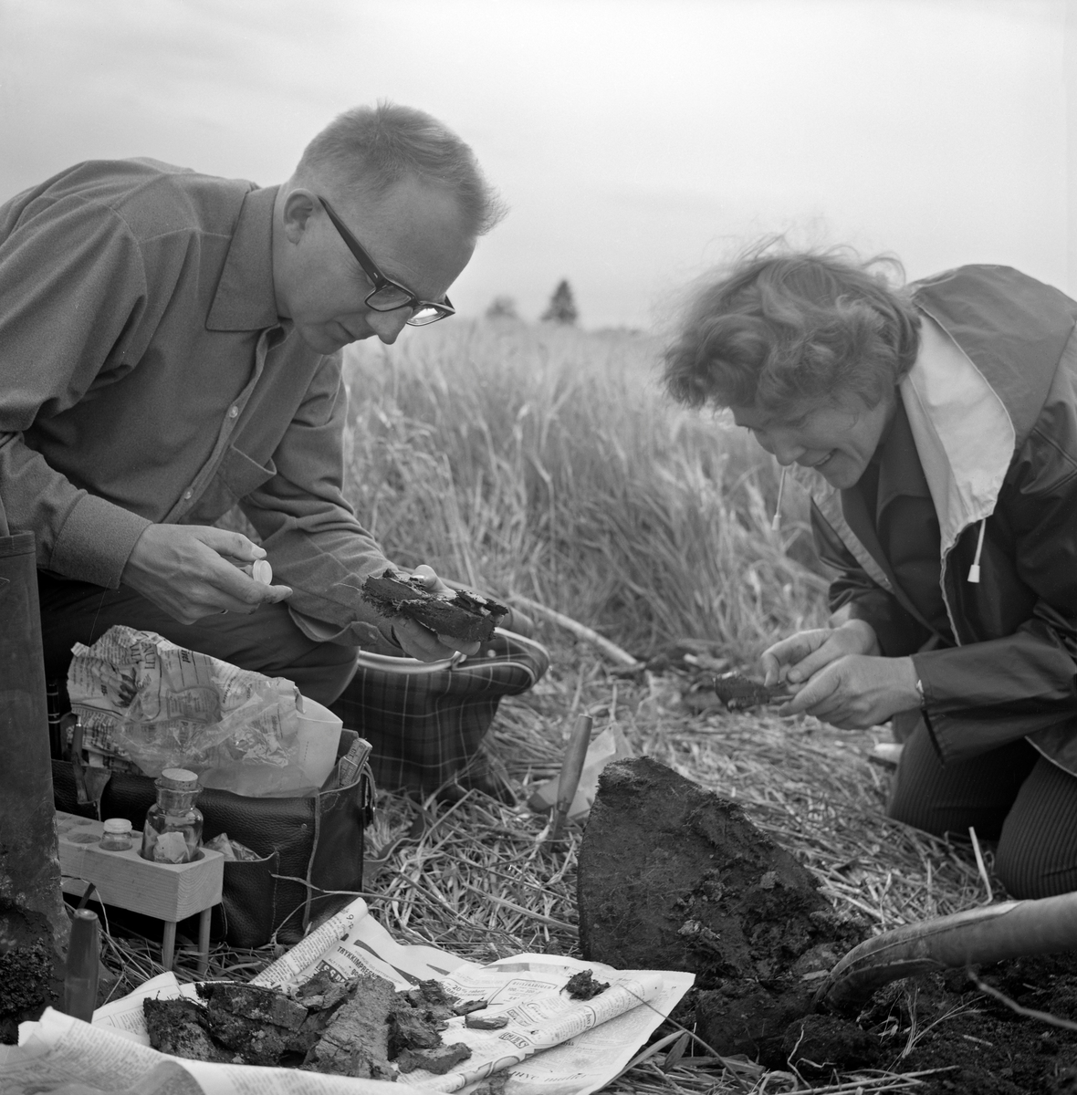 Utgravninger på garden Hov i Løten, Hedmark i 1966. Fotografiet er tatt under en naturhistorisk oppfølgingsundersøkelse som ble gjort etter at Gunnar Søberg i 1965 hadde funnet et elggevir påfallende langt nede i bakken under graving av ny trasé for kloakkledningen som gikk under et av jordene på Hov (jfr. SJF.01606 i museets gjenstandsdatabase). Pollenanalytikeren Kari Henningsmoen og limnologen Jan Økland gjorde en stratigrafisk undersøkelse av jordsmonnet for å kartlegge biologiske forhold på funnstedet gjennom årtusener. Her satt de ved kanten av undersøkelsessjakta som ble gravd et par meter fra funnstedet med feltutstyret sitt og prøver de hadde hentet opp fra sjakta. I bakgrunnen ser vi den frodige byggåkeren på Hov. Mer informasjon om dette prosjektet finnes under fanen «Opplysninger» og i de artiklene som er nevnt som litteraturreferanser til dette fotografiet.