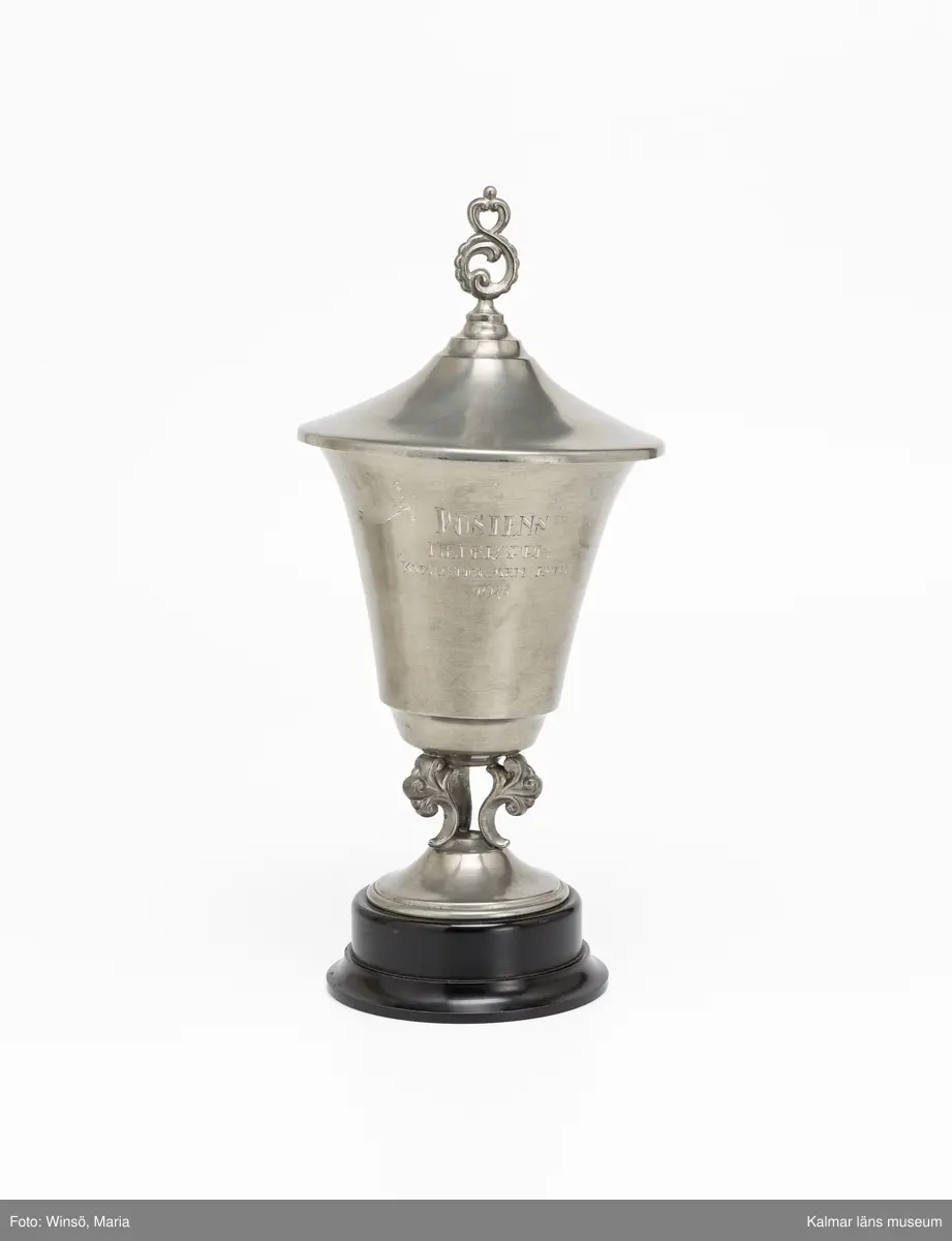 KLM 46137:3:1-2. Pokal, prispokal, lockpokal, metall, trä, färg. Prispokal i rokokostil med skål och fot i tenn (:3:1). Lock med dekor i form av bladrankor på toppen (:3:2). Skålen och foten binds samman av genombruten dekor i form av bladrankor. Foten är fastskruvad på svartmålad svarvad sockel av trä. Inskrift: POSTENS HEDERSPRIS "KVARNHOLMEN RUNT" 1941. Stämpel (insida lock): ortsstämpel för Stockholm SVENSKT TENN  N8.