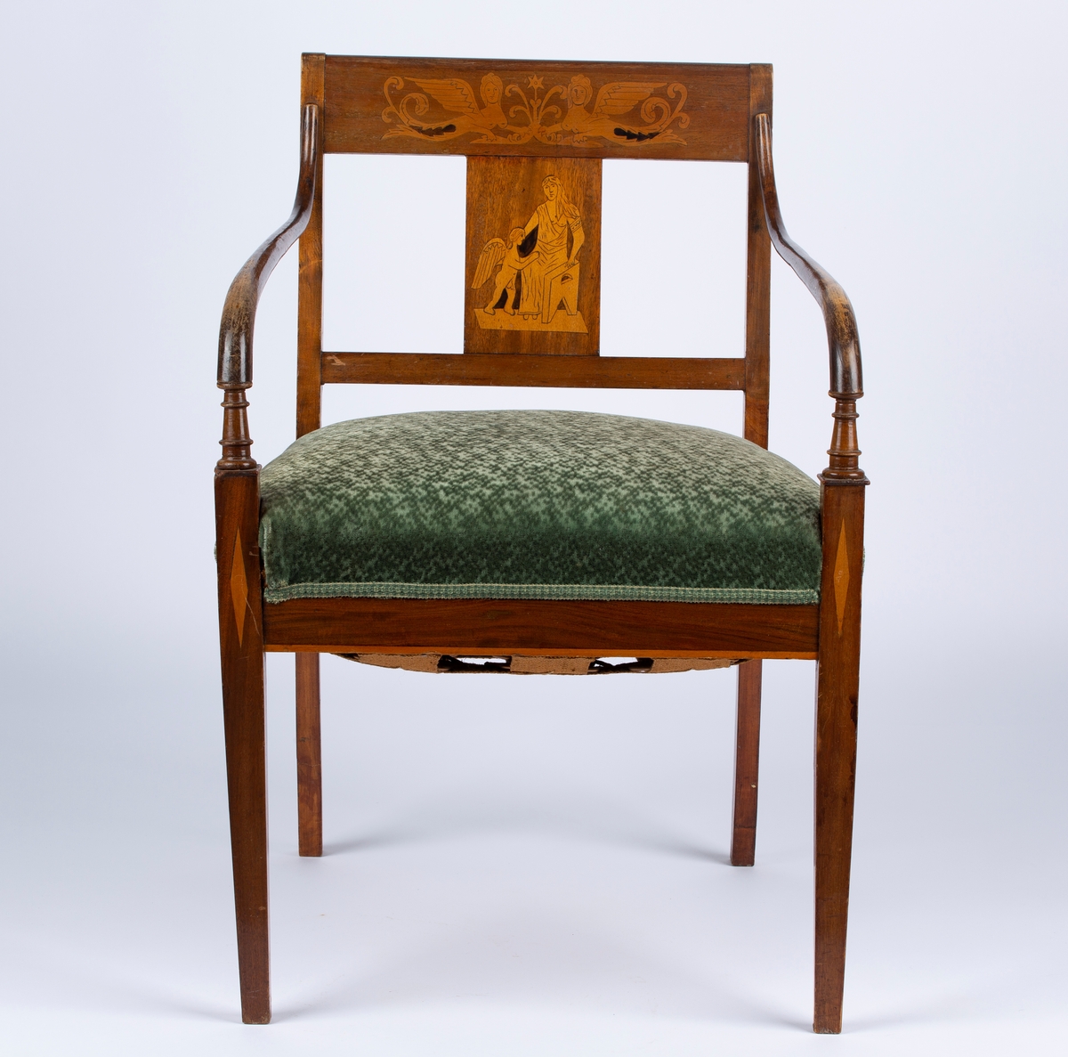 Sen-empire stol med armlener. Armlenene er mørkere enn resten av stolen. Trukket og stoppet sete. Svakt buede bakbein. Intarsia i tverrstykke og ryggbrett, klassiske figurmotiv og fabeldyr. Antikk-inspirert. Samme motiv på alle stolene, men med små ulikheter. Også samme motiv på skapene til sofaen, men ser ikke ut til å være samme kunstner. Enkel intarsia i de to fremste stolbenene, rutemønster, og en stripe i listen under stolsetet. Del av møblement.