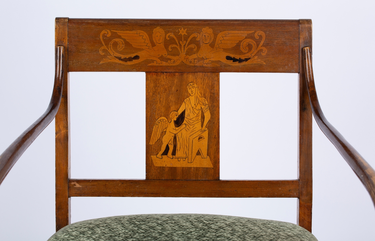Sen-empire stol med armlener. Armlenene er mørkere enn resten av stolen. Trukket og stoppet sete. Svakt buede bakbein. Intarsia i tverrstykke og ryggbrett, klassiske figurmotiv og fabeldyr. Antikk-inspirert. Samme motiv på alle stolene, men med små ulikheter. Også samme motiv på skapene til sofaen, men ser ikke ut til å være samme kunstner. Enkel intarsia i de to fremste stolbenene, rutemønster, og en stripe i listen under stolsetet. Del av møblement.