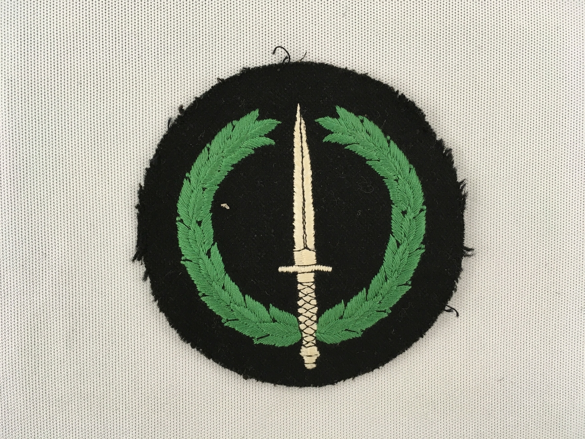 Motiv i kvitt og grønt  påsydd med maskin. Ein dolk omkransa av ein laurbærkrans