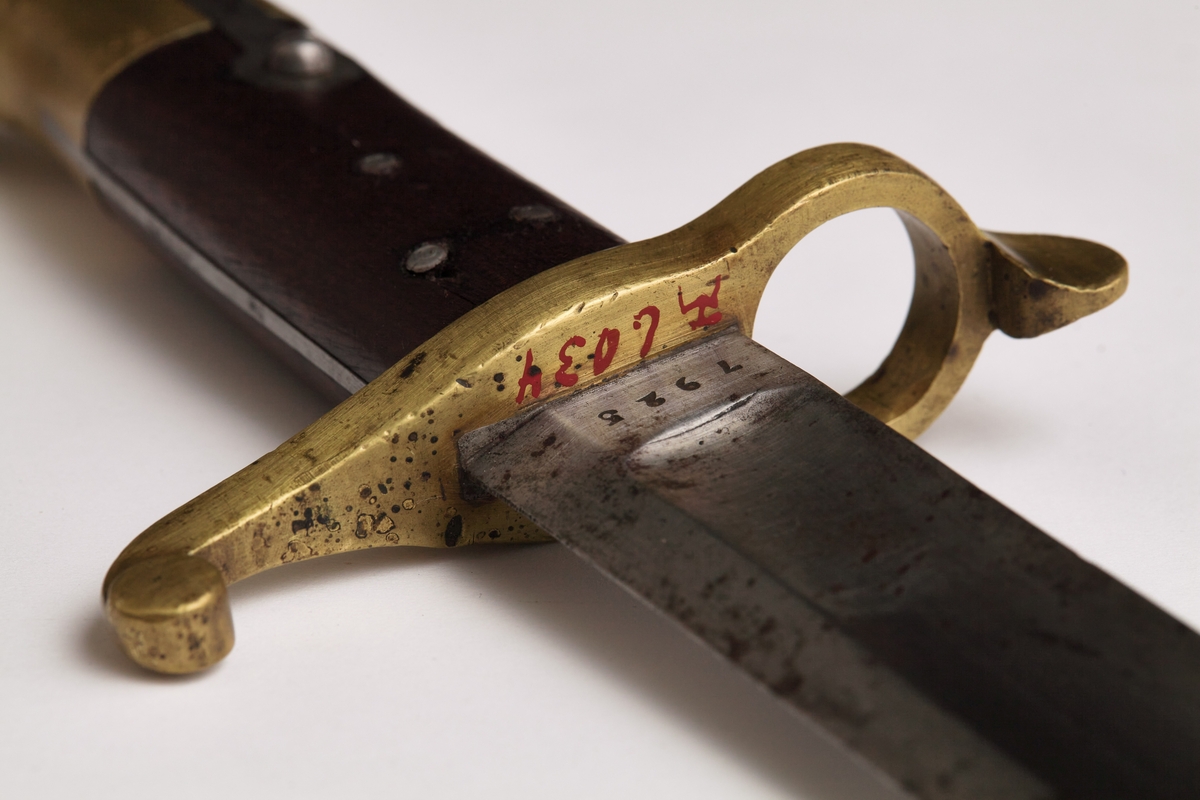 Sabelbajonett med jatangformet klinge, hulslipt, med tveegget spiss. Parerstang av messing, skjeftet av tre med en øvre del av messing. Parerstangens bakre arm danner en ring med diameter 23mm. Skjeftets øvre messingdel har spor for pipens bajonettklakk, låsingen skjer ved fjær og knapp av stål.
