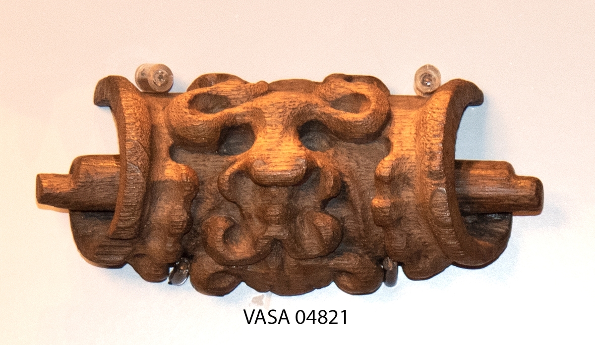 Rullverksornament.
Skulpterat litet, spänneliknande ornament, dekorerat i broskverksstil.
Det långsmala ornamentet är format som ett stycke kraftigt upprullad bit näver eller läder, med konkav bak- eller undersida. På baksidan är ornamentet profilerat för en list. Broskdekoren på ovansidan är formad som en maskaron.
Ornamentet är mycket välbevarat.

Text in English: A small, buckle-like ornament, decorated in a style similar to earshell work.
The long narrow ornament is formed as a piece of tightly rolled bark or leather, with a concave back or underside so as to match a batten or the like. The reverse side is profiled to fit a moulding. The earshell work on the upper side is shaped as a mask.
The ornament is well preserved.