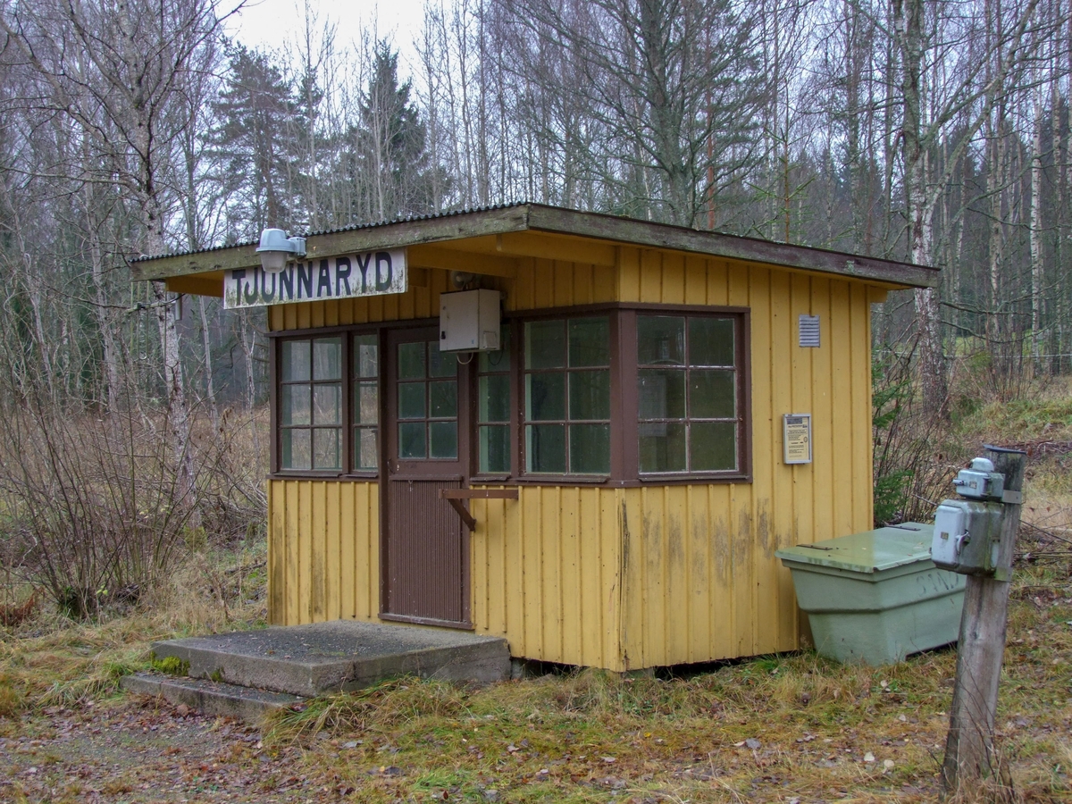 Hållplatskur från Tjunnaryd. Målad i ljusgul kulör med ljusgrå dörr, dörrfoder och fönsterfoder. Spröjsade fönster utmed hela framsidan, även på dörren, och delvis på sidorna.