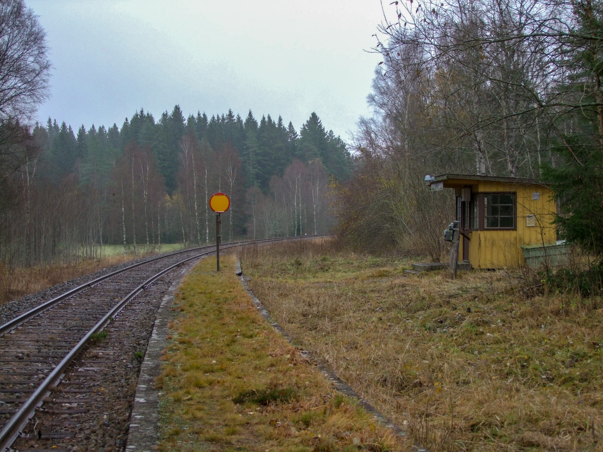 Hållplatskur från Tjunnaryd. Målad i ljusgul kulör med ljusgrå dörr, dörrfoder och fönsterfoder. Spröjsade fönster utmed hela framsidan, även på dörren, och delvis på sidorna.