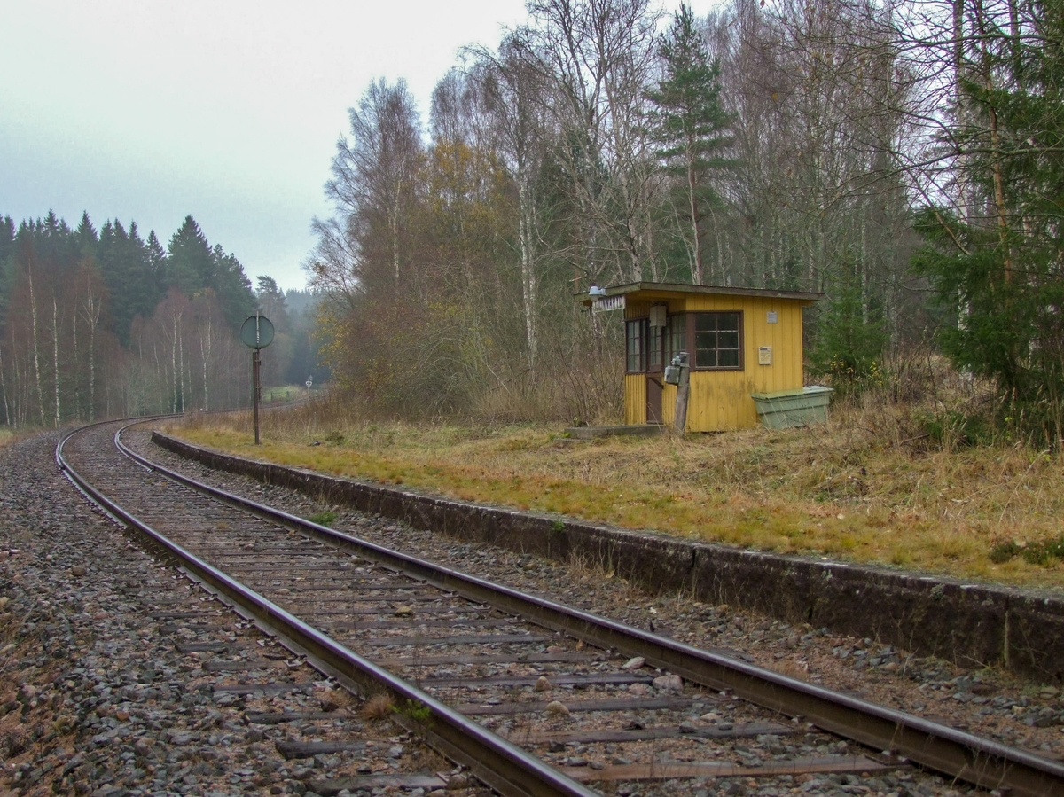 Hållplatskur från Tjunnaryd. Målad i ljusgul kulör med ljusgrå dörr, dörrfoder och fönsterfoder. Spröjsade fönster utmed hela framsidan, även på dörren, och delvis på sidorna.