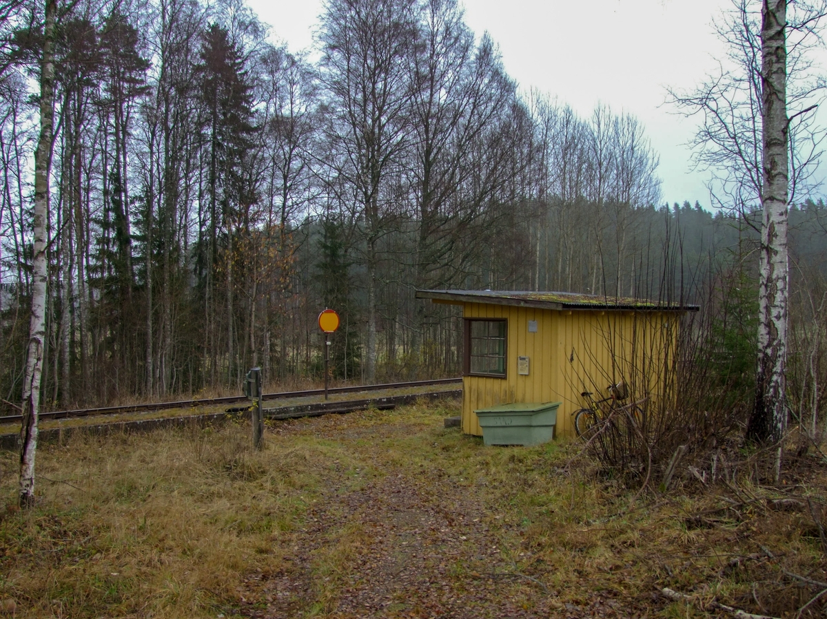 Hållplatskur från Tjunnaryd. Målad i ljusgul kulör med ljusgrå dörr, dörrfoder och fönsterfoder. Spröjsade fönster utmed hela framsidan, även på dörren, och delvis på sidorna.