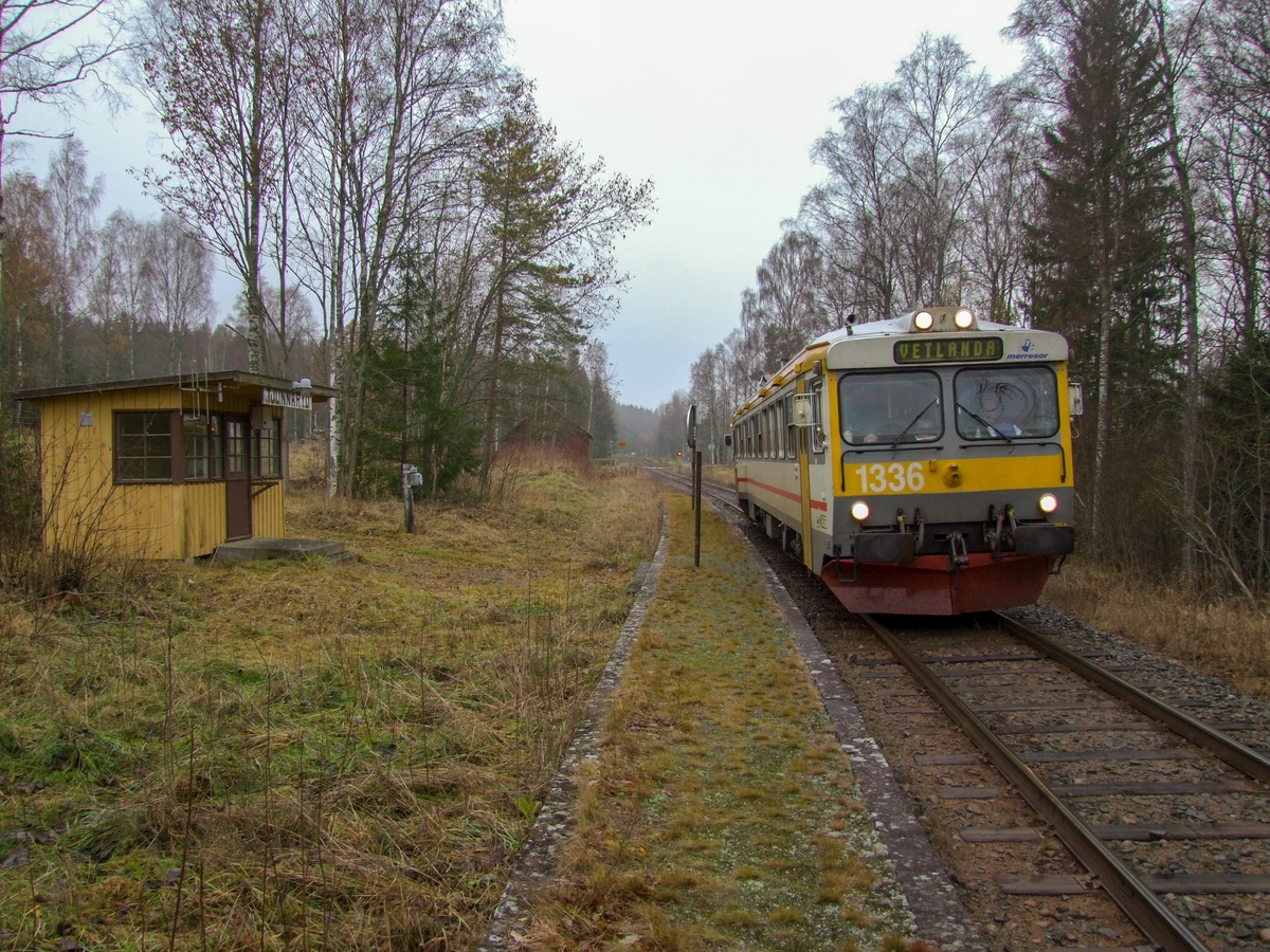 Hållplatskur från Tjunnaryd. Målad i ljusgul kulör med ljusgrå dörr, dörrfoder och fönsterfoder. Spröjsade fönster utmed hela framsidan, även på dörren, och delvis på sidorna.