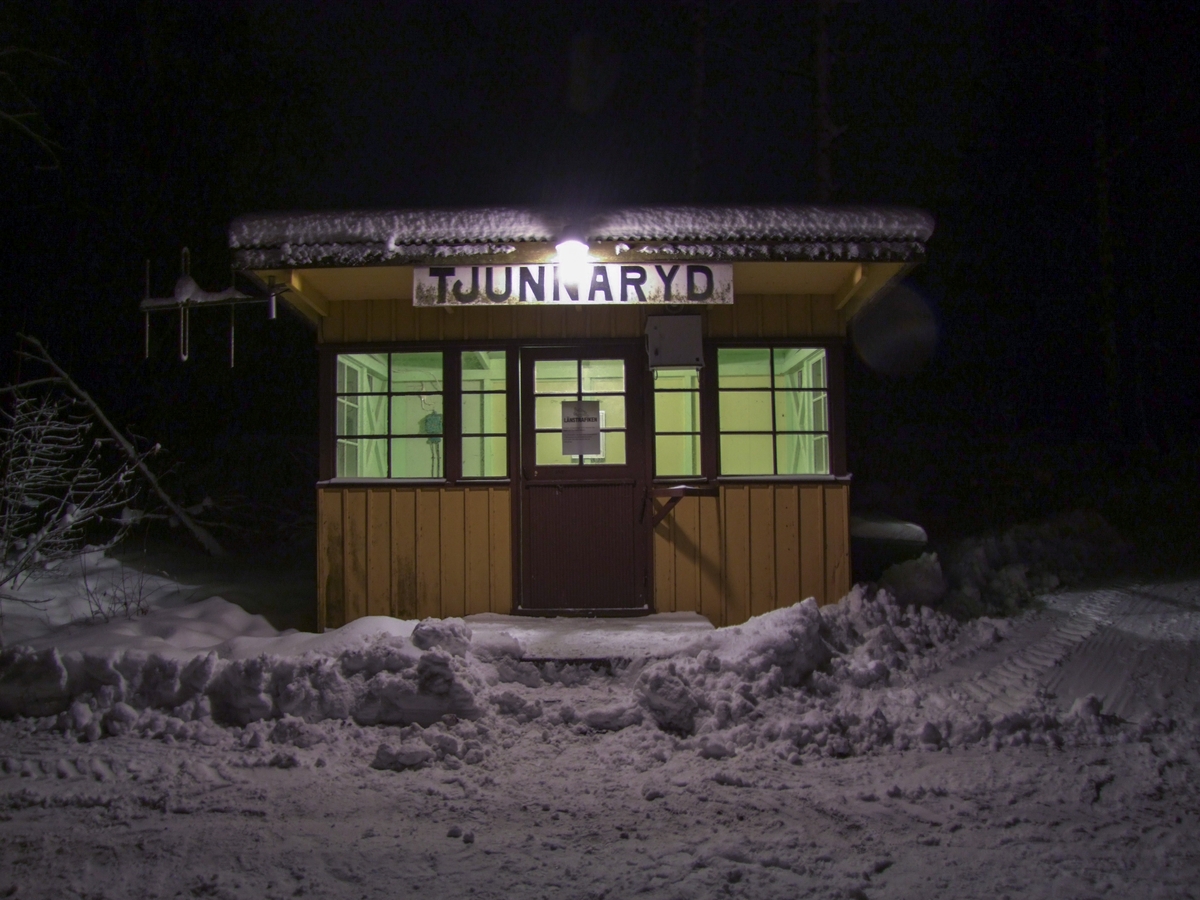 Hållplatskur från Tjunnaryd. Målad i ljusgul kulör med ljusgrå dörr, dörrfoder och fönsterfoder. Spröjsade fönster utmed hela framsidan, även på dörren, och delvis på sidorna.