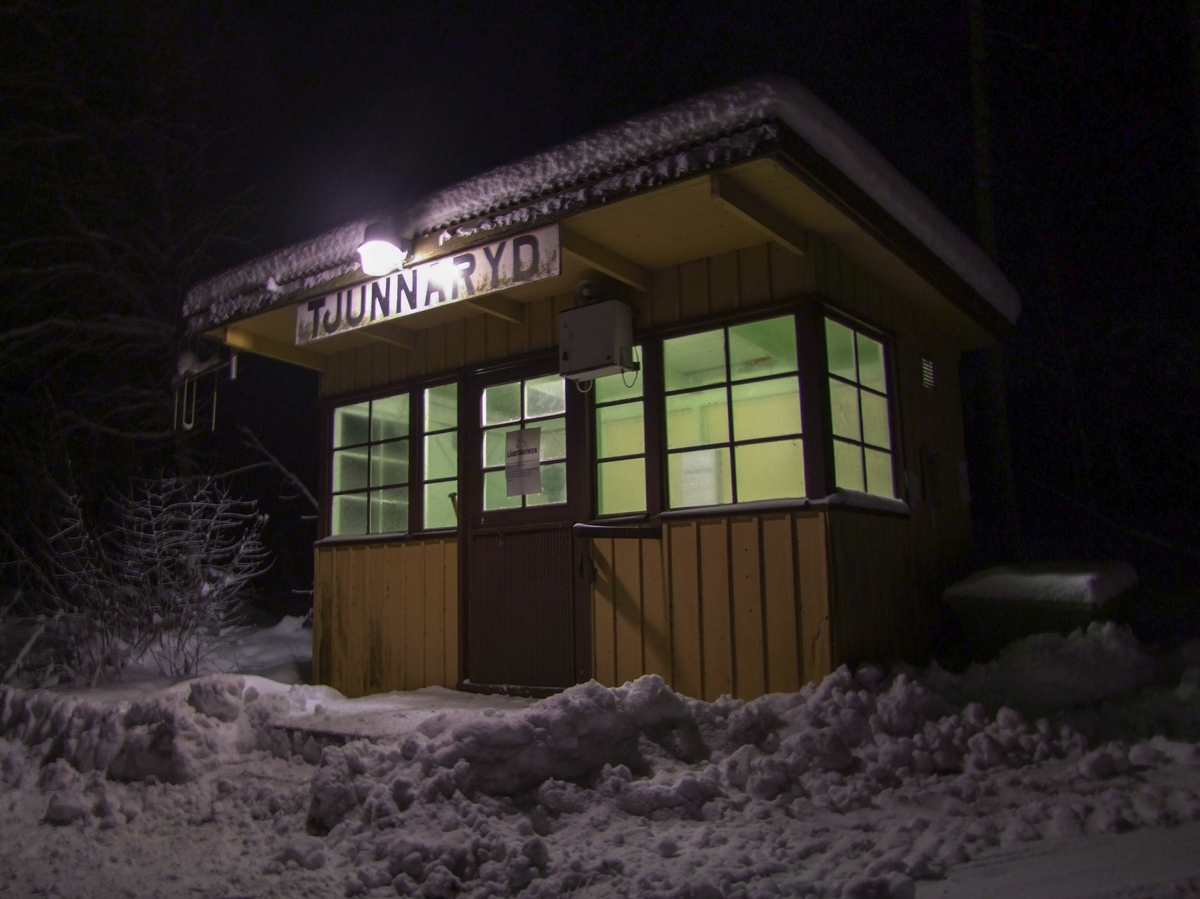 Hållplatskur från Tjunnaryd. Målad i ljusgul kulör med ljusgrå dörr, dörrfoder och fönsterfoder. Spröjsade fönster utmed hela framsidan, även på dörren, och delvis på sidorna.