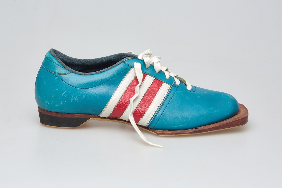 En skisko av turkisblått lær med røde og hvite striper på siden. Indre tunge og snøring med hvite skolisser. Beige såle av gummi.