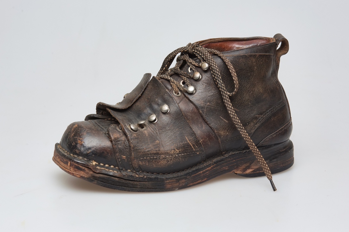 En skisko av brunt lær. Indre tunge og snøring gjennom maljer og hekter med skolisser. Overlappende lag med snøring utenpå dette, med hekter. Såle av lær.